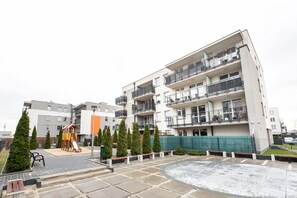 Un immeuble résidentiel à plusieurs étages, doté de balcons, d’une aire de jeux et d’une cour dallée.