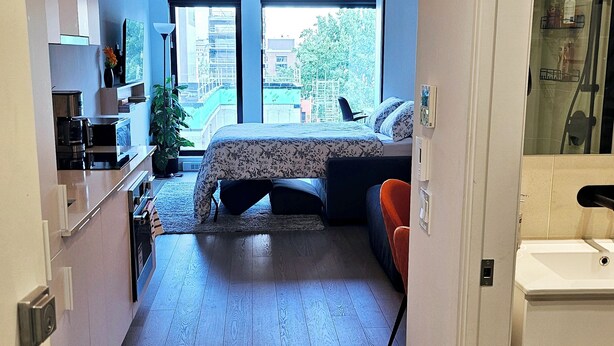 A narrow hallway leading to a bedroom with a bed, a small seating area, and a kitchenette.