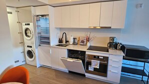A modern kitchen with white cabinets, a built-in dishwasher, a microwave, and a coffee maker. There is a dining area with a table and orange chairs.