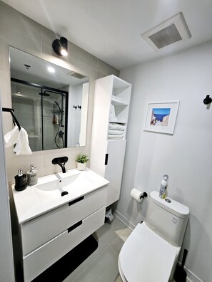 Un baño moderno con lavamanos, espejo y retrete blancos. Incluye un box de ducha y un perchero para toallas.