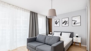 Ein modernes Schlafzimmer mit grauem Sofa, einem Bett mit weißer Bettwäsche, einem Nachttisch mit Lampe und abstrakter Wandkunst.