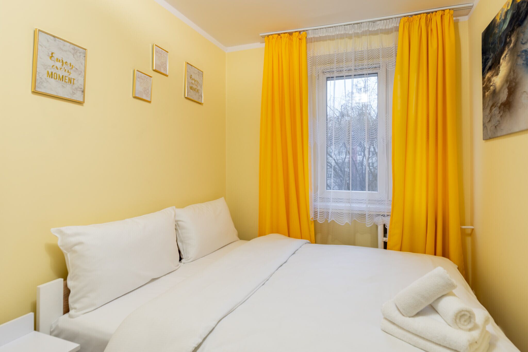 Hotellihuone, jossa on sänky, keltaiset verhot, pitsiverhoilla varustettu ikkuna ja seinällä olevia kehystettyjä tauluja.