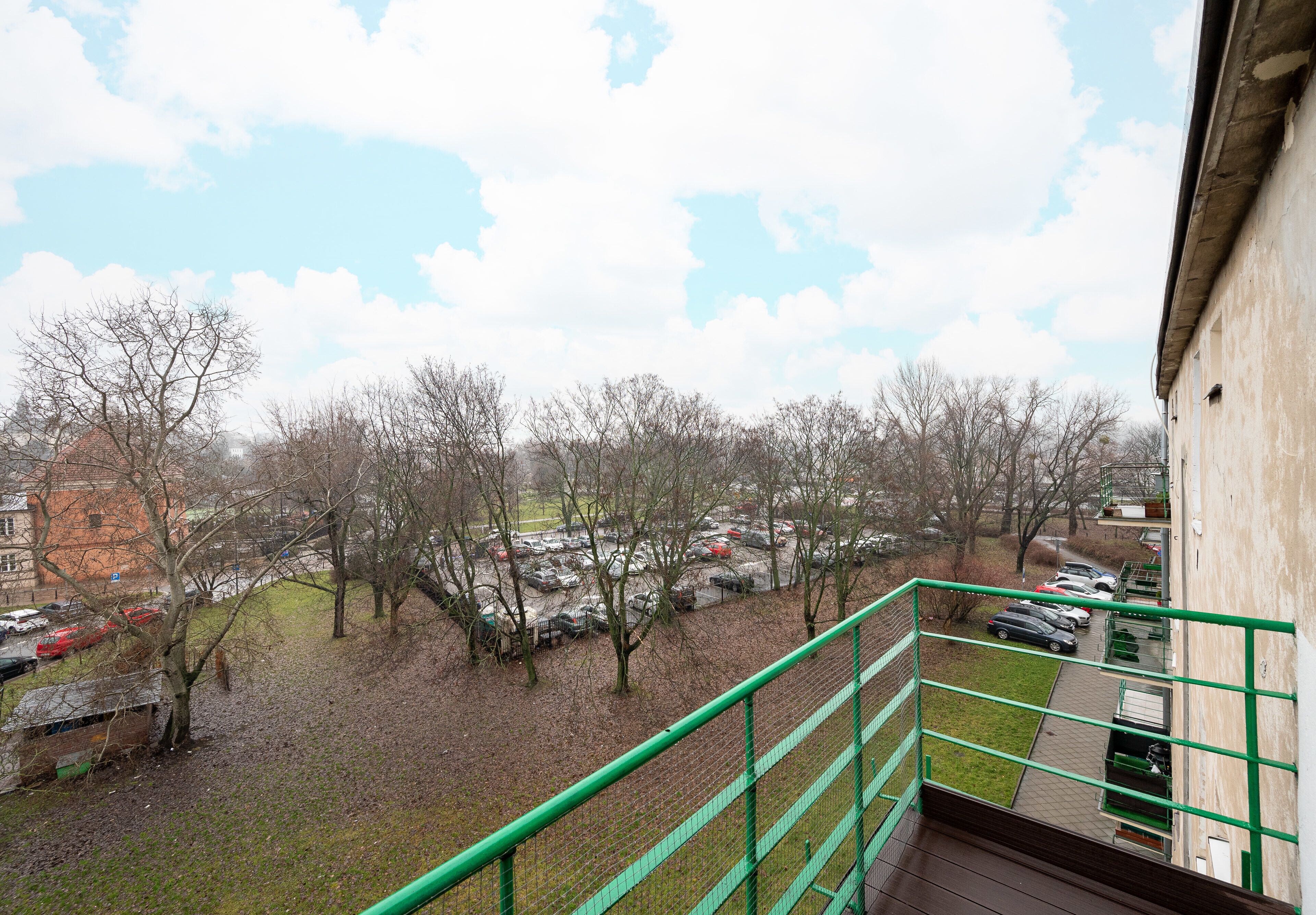 A view from a balcony overlooking a parking area with trees and a building in the background.