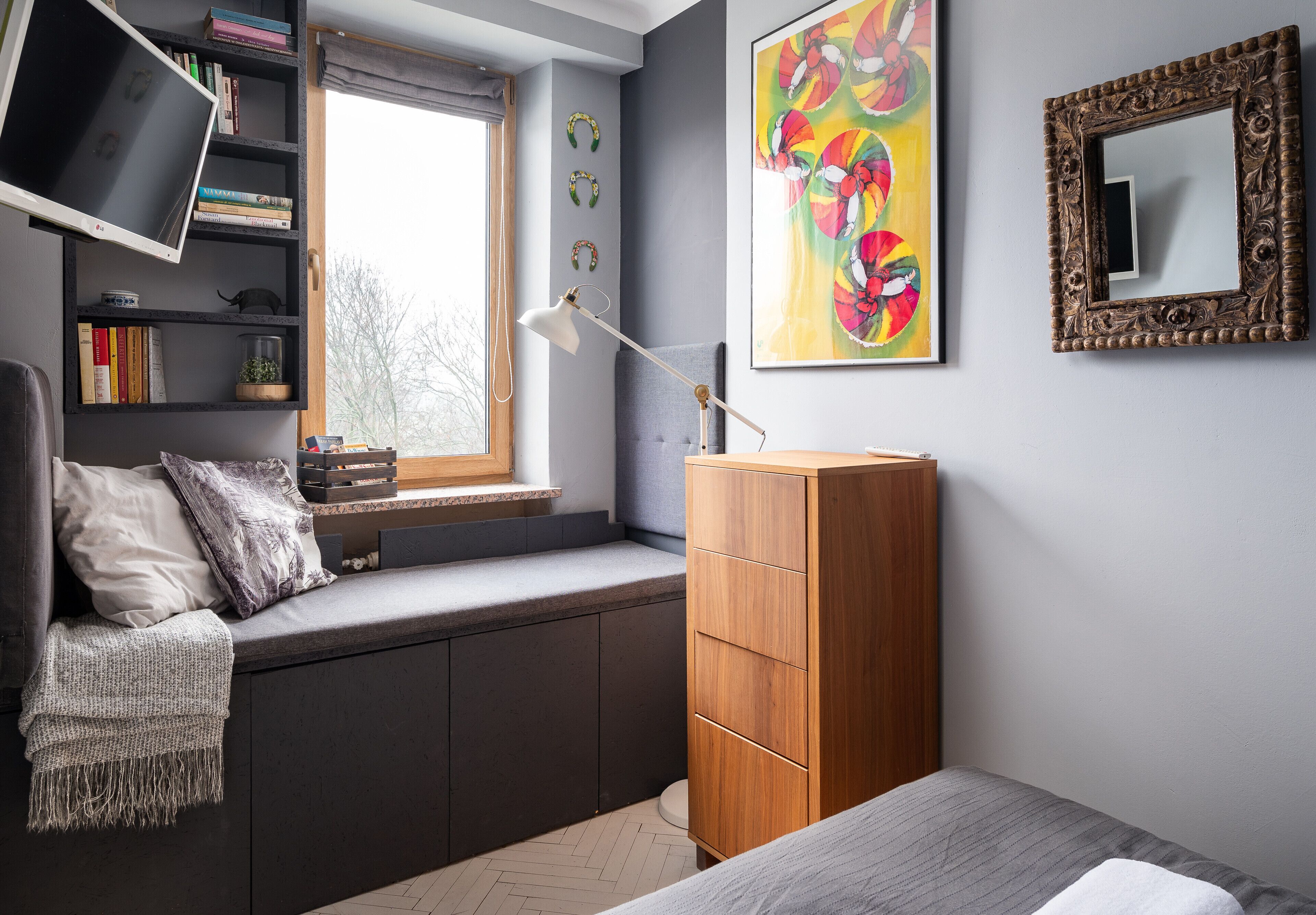 Une chambre avec une banquette intégrée, une armoire en bois, un bureau avec une lampe, un téléviseur et un grand tableau.