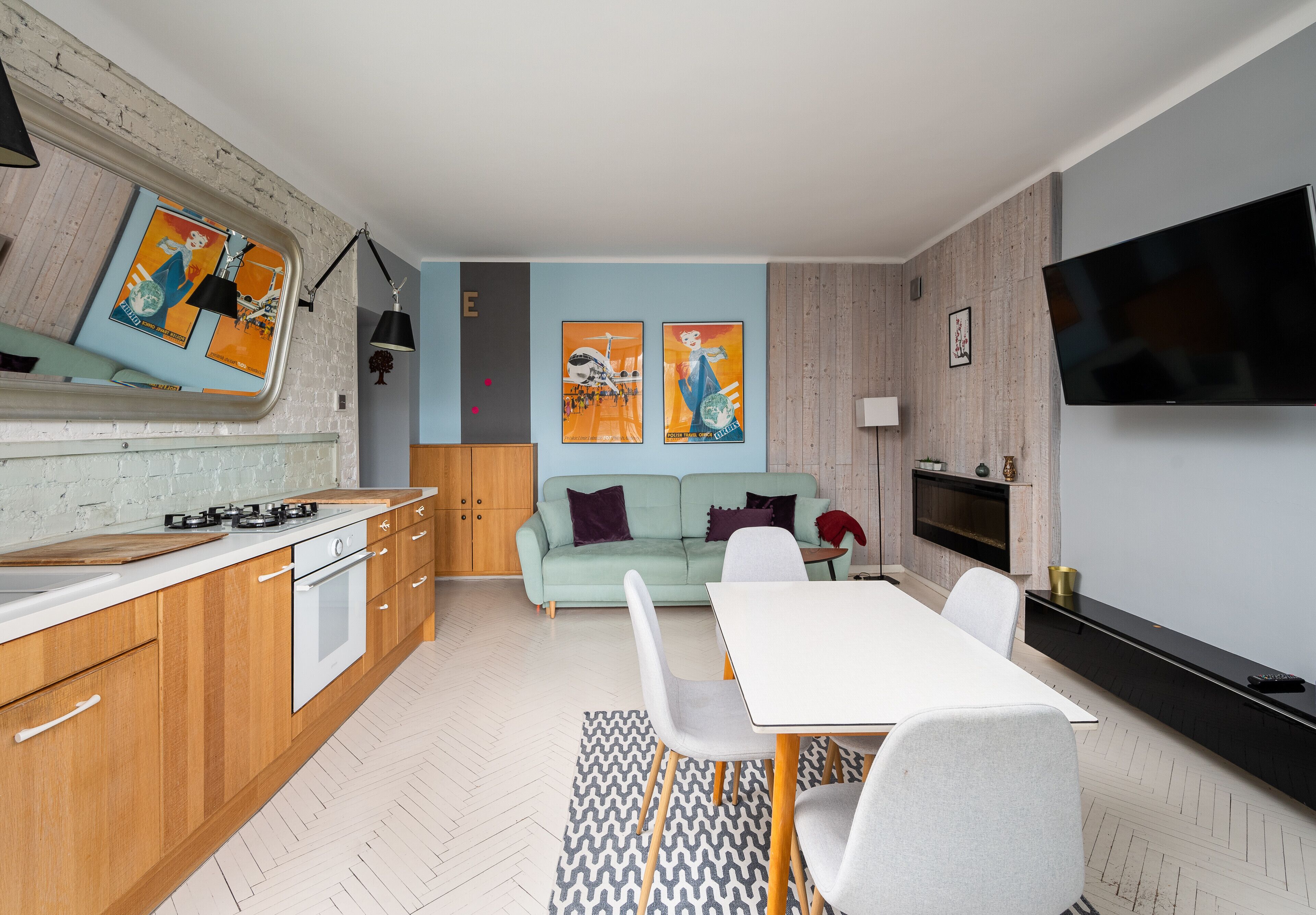 A modern kitchen with wooden cabinets, a white countertop, and a built-in oven. There is a dining area with a white table and chairs, and a sofa in the background.