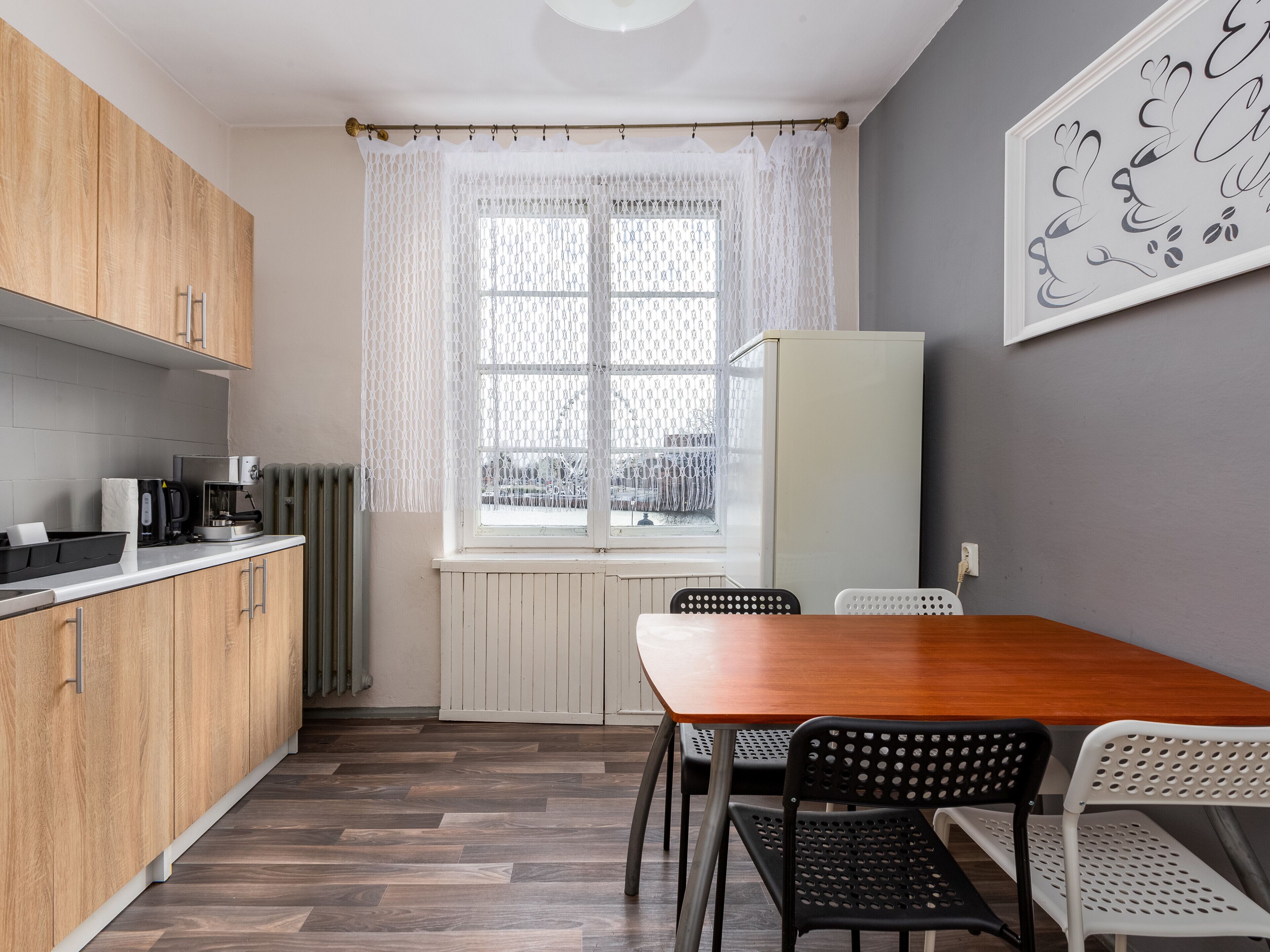 Et lille køkken med træskabe, et hvidt køleskab, et spisebord med to stole og et vindue med et mønstret gardin.