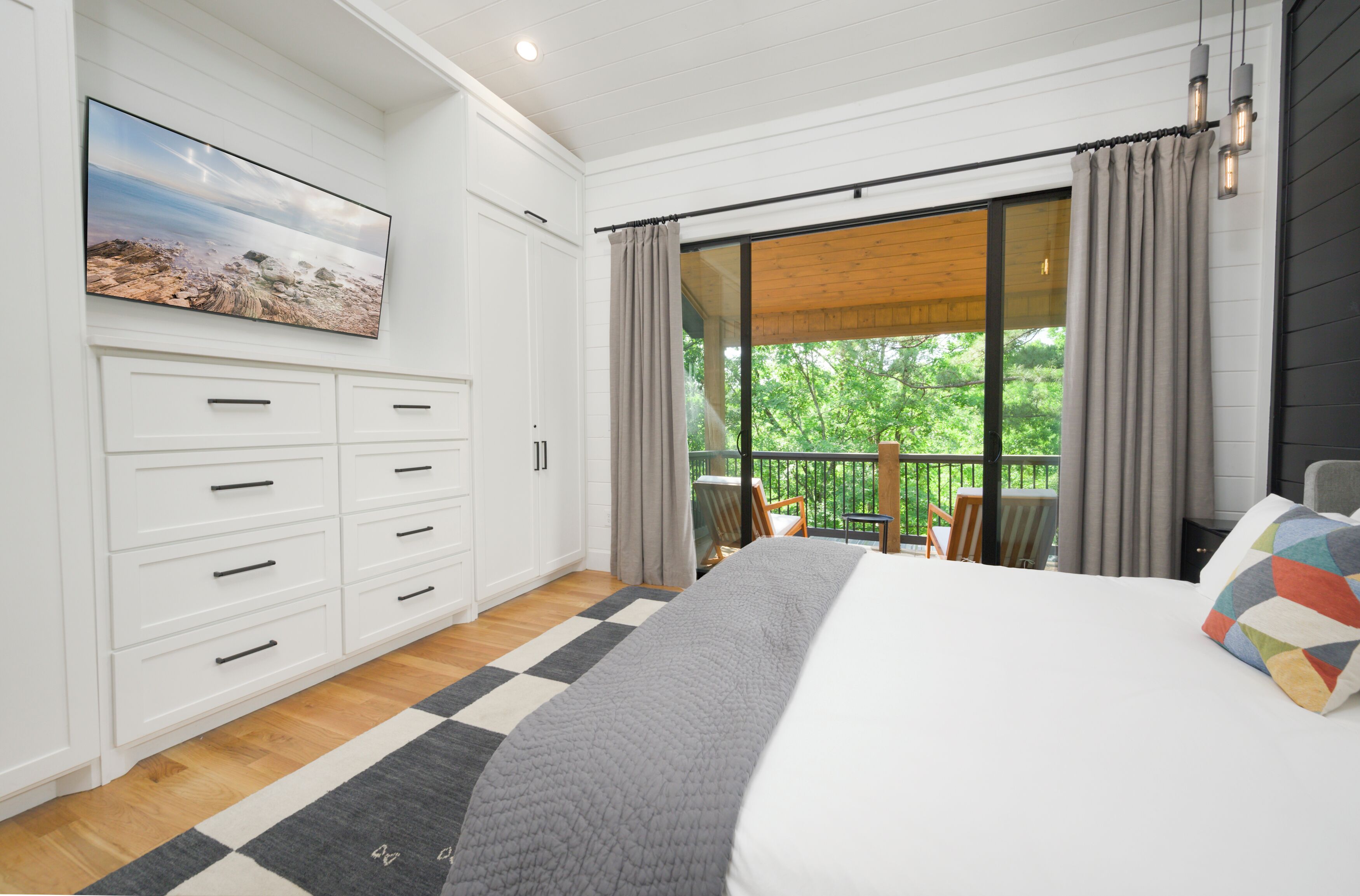 A bedroom with a large bed, a dresser, a painting, and a balcony with a view of greenery.