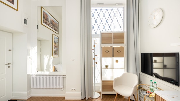 A modern hotel room with a white color scheme, a large window with a view, a white chair, a TV, and a radiator.