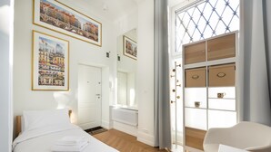 A hotel room with a bed, a chair, a window with curtains, and two framed pictures on the wall.