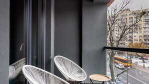 Un balcón con dos sillas blancas de mimbre y una pequeña mesa redonda, con vista a una calle de la ciudad con edificios y automóviles estacionados.