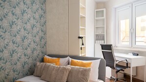 A neatly made bed with a yellow headboard, a grey and yellow pillow arrangement, a white desk with a chair, and a window with blinds. - Park Praski 2-Bedroom Apartment (Warszawa)