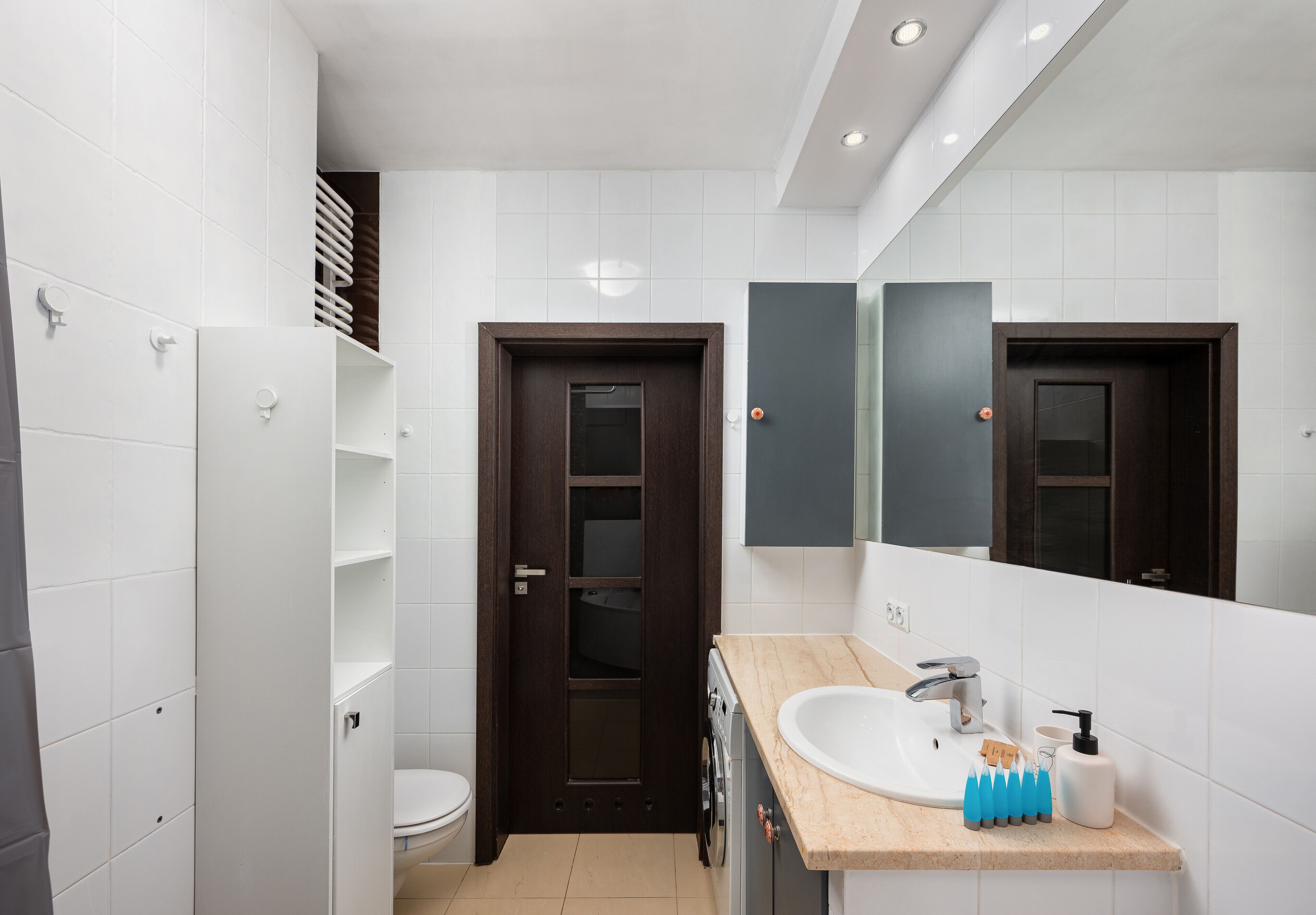 Un baño moderno con baldosas blancas, una puerta de madera oscura, un lavamanos con espejo y un perchero para toallas.
