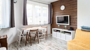 A room with a wooden wall, a TV, a white cabinet, a yellow chair, and a table with chairs. - Park Praski 2-Bedroom Apartment (Warszawa)
