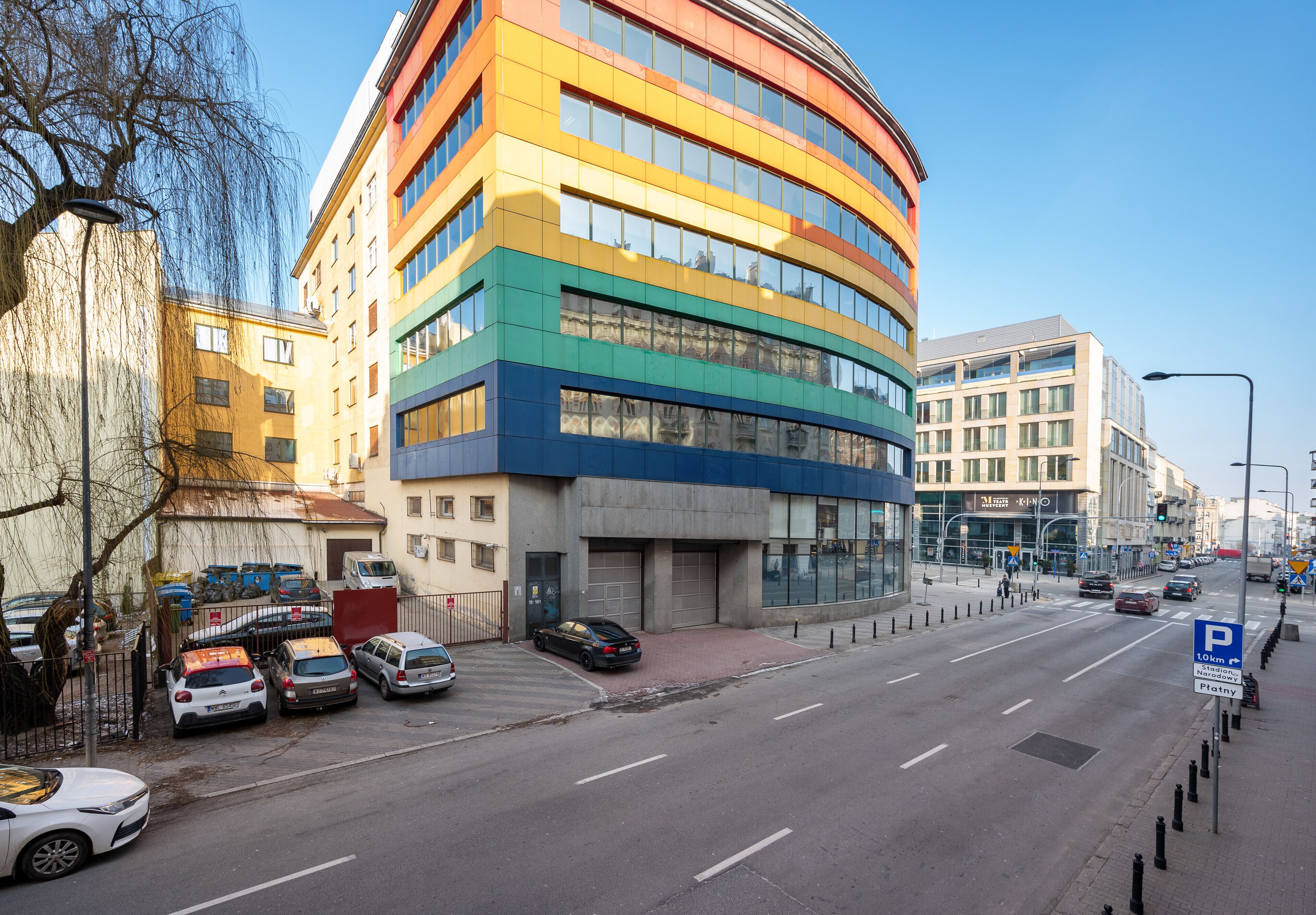 A multi-story building with a curved facade, parked cars along the street, and a clear blue sky.