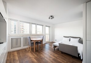 Ein modernes, helles Zimmer mit Holzboden, einem Esstisch mit Stühlen, einem Bett mit grauem Kopfteil und großen Fenstern mit Blick auf Gebäude.