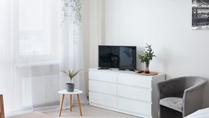 Un salon moderne avec un téléviseur à écran plat posé sur une commode blanche, un fauteuil gris, une petite table ronde agrémentée d’une plante en pot et une table à manger en bois avec un chemin de table à motifs floraux.