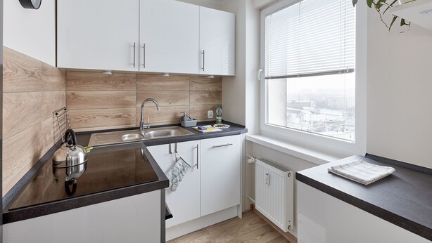 Une cuisine moderne avec des meubles blancs, une bouilloire en acier inoxydable et une fenêtre équipée de stores.