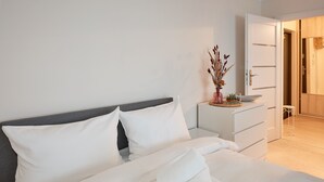 Un lit bien fait, avec du linge de lit blanc et une serviette pliée, une table de chevet ornée d’un vase de fleurs séchées et une commode blanche.