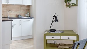 Une cuisine compacte avec des meubles blancs, un pare-feu en bois et un évier. On y trouve un bureau vert avec une chaise, un miroir et une lampe.