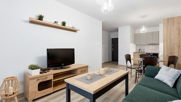 A modern living room with a flat-screen TV, wooden entertainment unit, green sofa, and a dining area with a table and chairs. - Poznań Airport Comfy Apartment (Poznań)