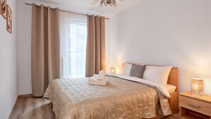 A neatly made bed with a beige quilt, a wooden nightstand with a lamp, and a window with curtains. - Poznań Airport Comfy Apartment (Poznań)