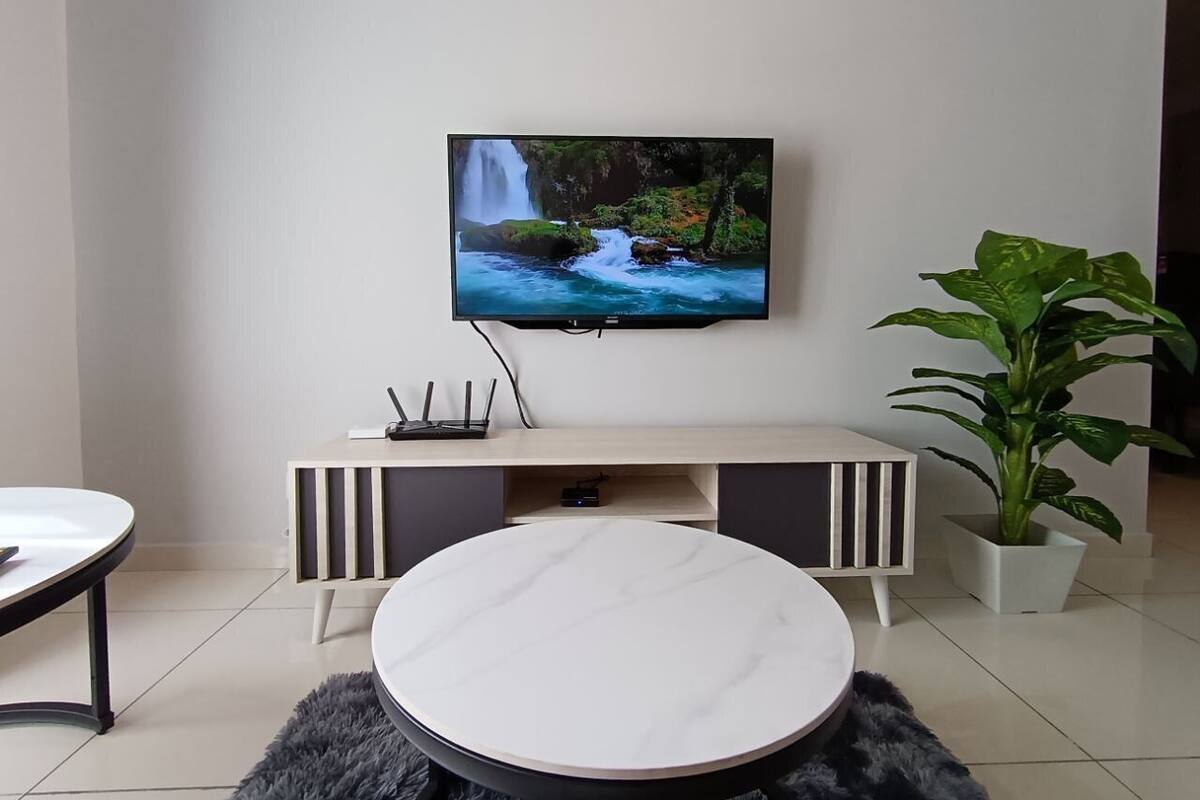Een moderne woonkamer met een flatscreen televisie aan de muur, een ronde salontafel, een witte bank, een kleine witte tafel en een potplant.