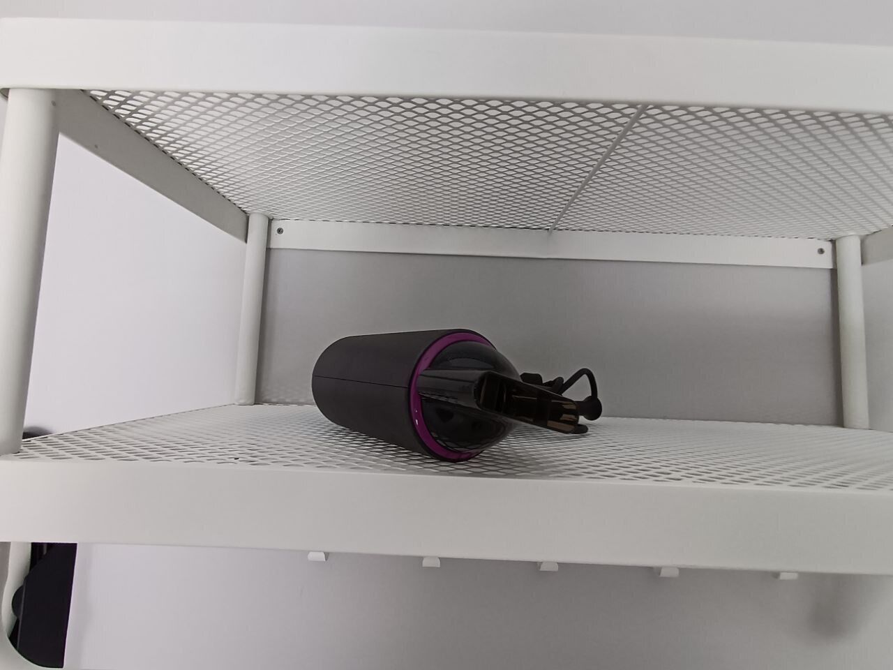 Een wit metalen schap met een gaasbovenkant en een zwart cilindrisch voorwerp met een paarse ring.