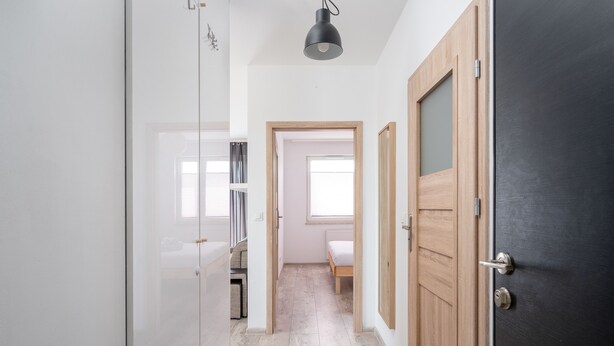 Un couloir étroit avec une porte en bois sur la droite, une porte blanche sur la gauche et un lit au fond.