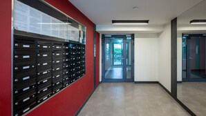 Un couloir avec une rangée de boîtes aux lettres sur le côté gauche, une porte au fond et un ascenseur en verre sur le côté droit.