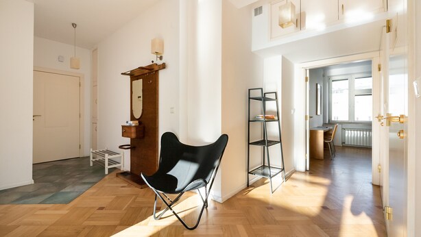 Ein geräumiges Zimmer mit einem Holzboden im Fischgrätmuster, einem schwarzen Stuhl, einem Holzschrank mit Spiegel und einem Regal.
