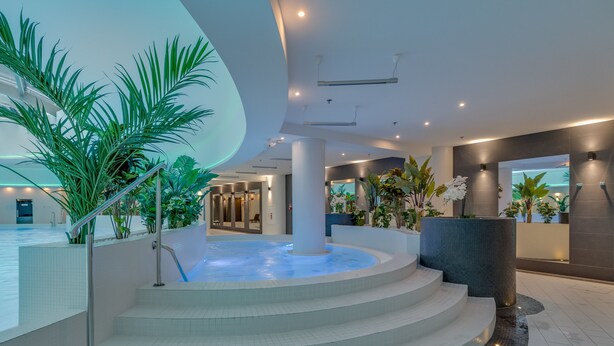 A modern indoor pool area with a central hot tub, surrounded by steps and various plants.