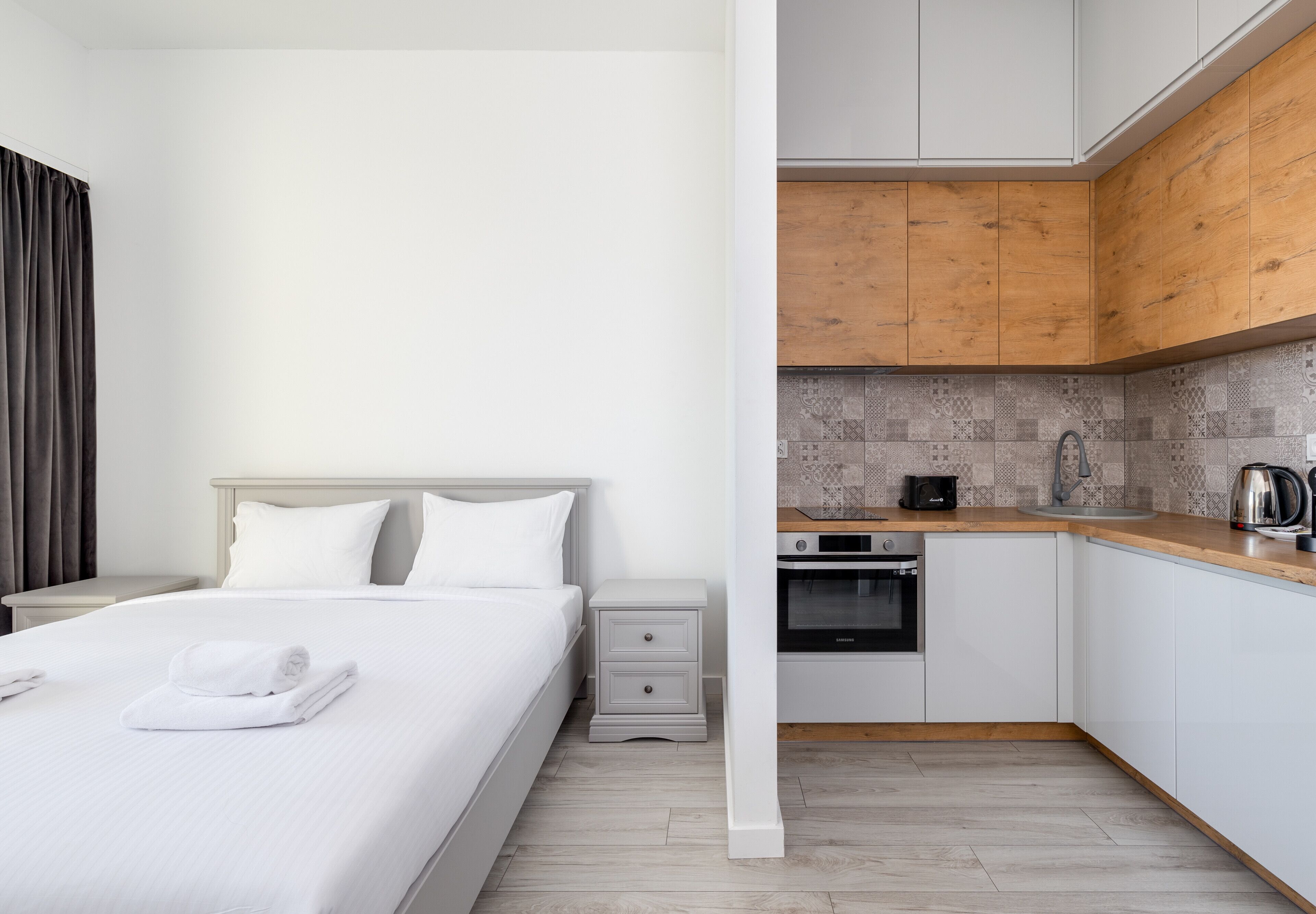 A hotel room with a bed, a nightstand, a small drawer, a kitchen area with wooden cabinets, a tiled backsplash, and a coffee maker.