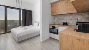 A modern hotel room with a bed, a kitchenette, and a sliding glass door to a balcony.