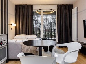 A modern hotel room with a bed, a round table, white chairs, a TV, and a window with curtains.