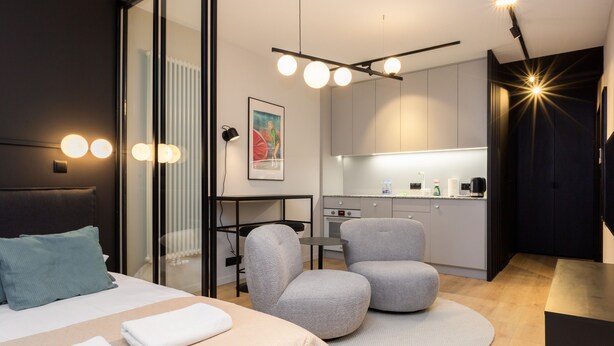 A modern hotel room with a bed, two armchairs, a small desk, and a kitchenette. - Quercus Deluxe Studio #B (Gdansk)