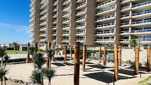 A high-rise building with multiple balconies, a sandy ground area with palm trees, and a clear blue sky. - Beautiful 3 bedroom/3 bathroom Condo In Las Palomas Beach & Golf Resort (Puerto Penasco)