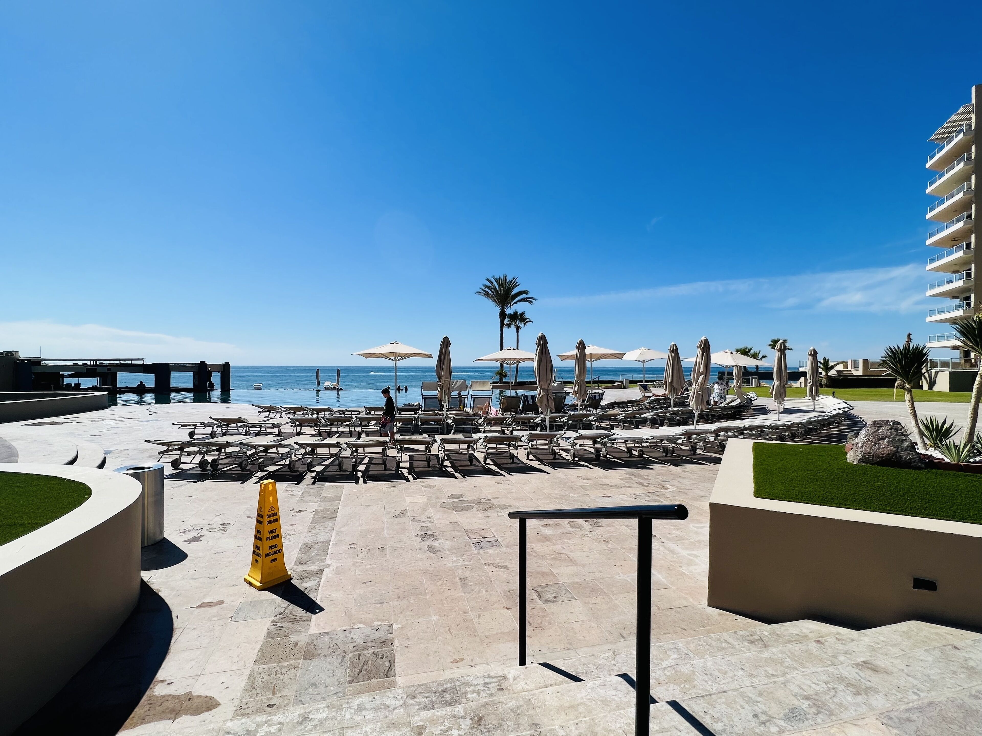 Área de piscina de um resort com espreguiçadeiras e guarda-sóis, céu azul límpido e vista para o oceano.