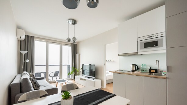 Un salon moderne avec un canapé, une table à manger et un téléviseur. On peut apercevoir une cuisine avec des meubles blancs et des appareils électroménagers.