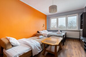 A room with two beds, a wooden table, a hanging light fixture, and a window with a view of trees.