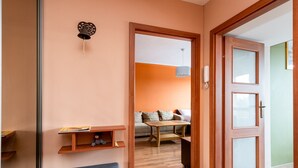 Habitació amb una banca de fusta, una petita estanteria plena de llibres, un campanar fixat a la paret i una porta que dona a una altra habitació amb un sofà i una taula.