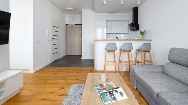 A modern living room with a grey sofa, wooden coffee table, and a magazine. There are two bar stools and a kitchen area with a black range hood.