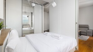 A modern hotel room with a large bed, a wooden desk, a grey sofa, and a view of the city through a large window.