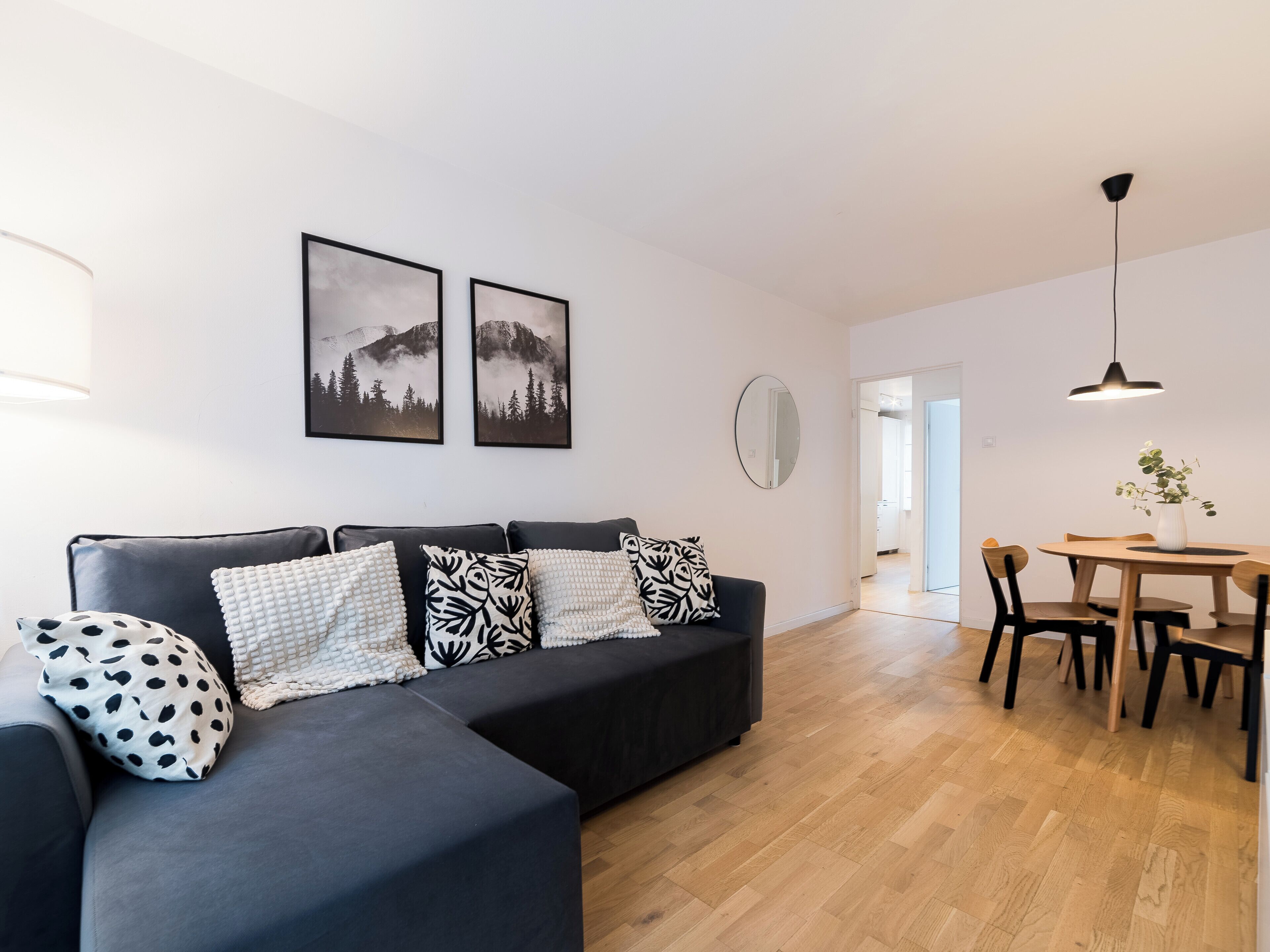 Un salon moderne avec un canapé d’angle gris foncé, deux coussins décoratifs noirs et blancs à motifs, une table de salle à manger ronde en bois avec quatre chaises et une suspension.