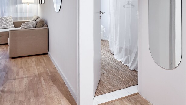 Una habitación moderna y luminosa con suelo de madera, un sofá color beige, un espejo redondo y una cama con dosel blanco.