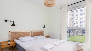 Une chambre à coucher comprenant un lit, des tables de chevet, une lampe, une fenêtre donnant sur des immeubles et un radiateur.
