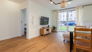 Un salon moderne avec un plancher en bois, une table à manger en bois, un téléviseur fixé au mur et une grande fenêtre donnant sur un immeuble et des voitures garées.