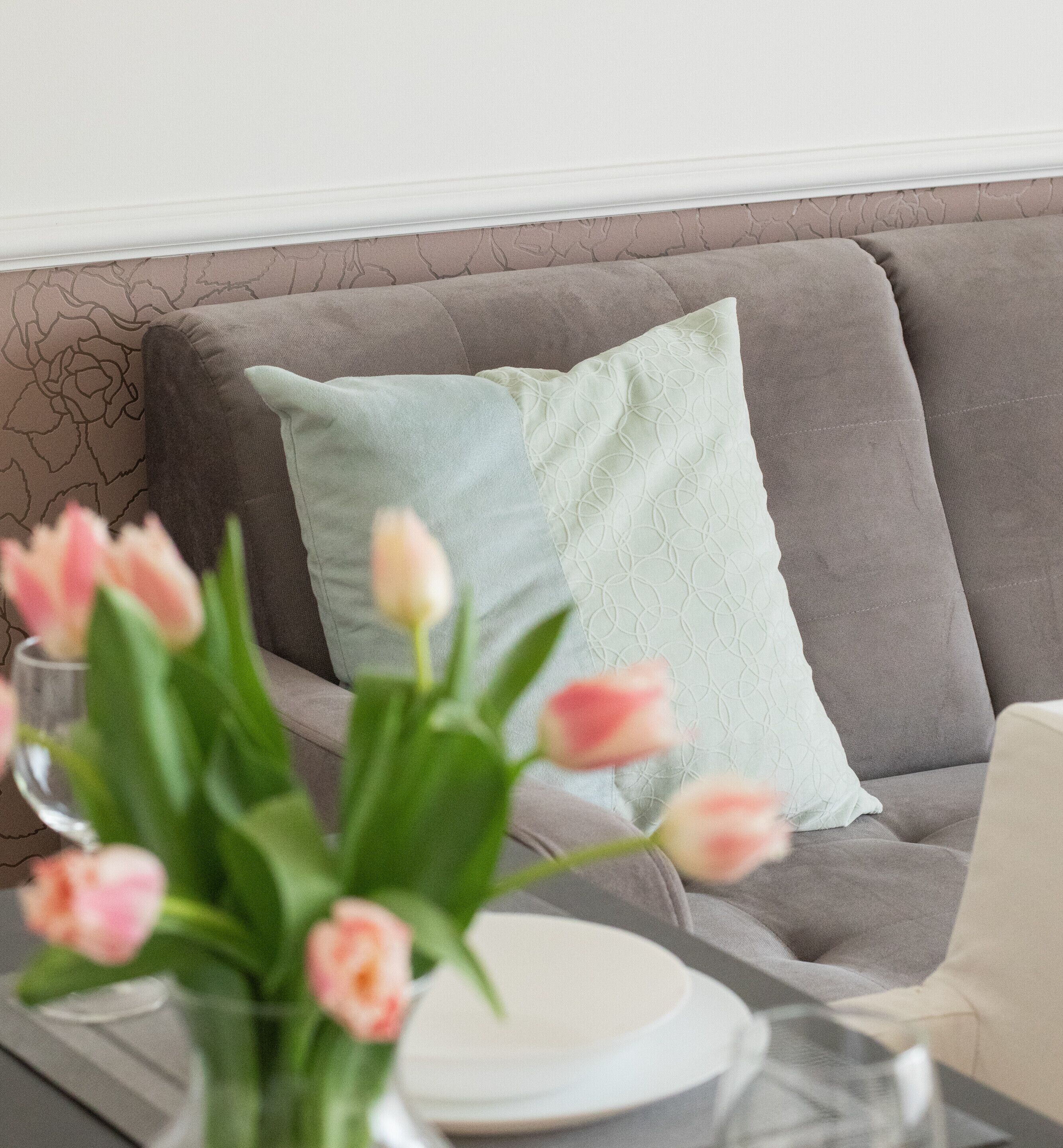 A modern living room with a grey sofa, a light green pillow, and a vase with pink tulips on a table.
