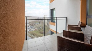 Een balkon met rieten meubilair, een glazen hekwerk en uitzicht op een parkeerplaats en gebouwen.