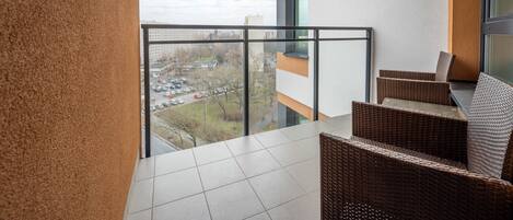 Ein Balkon mit Korbmöbeln, einer Glasreling und Blick auf einen Parkplatz sowie Gebäude.