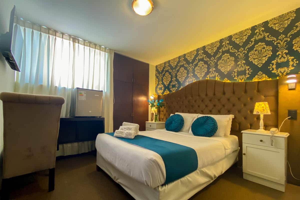 A hotel room with a large bed, a desk, a chair, a nightstand, and a wall with patterned wallpaper.